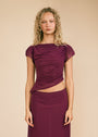 A woman modeling a two-piece outfit consisting of a deep purple wrap-style top with cap sleeves and a matching high-low purple skirt, paired with black pointed-toe heels, against a solid beige studio background.