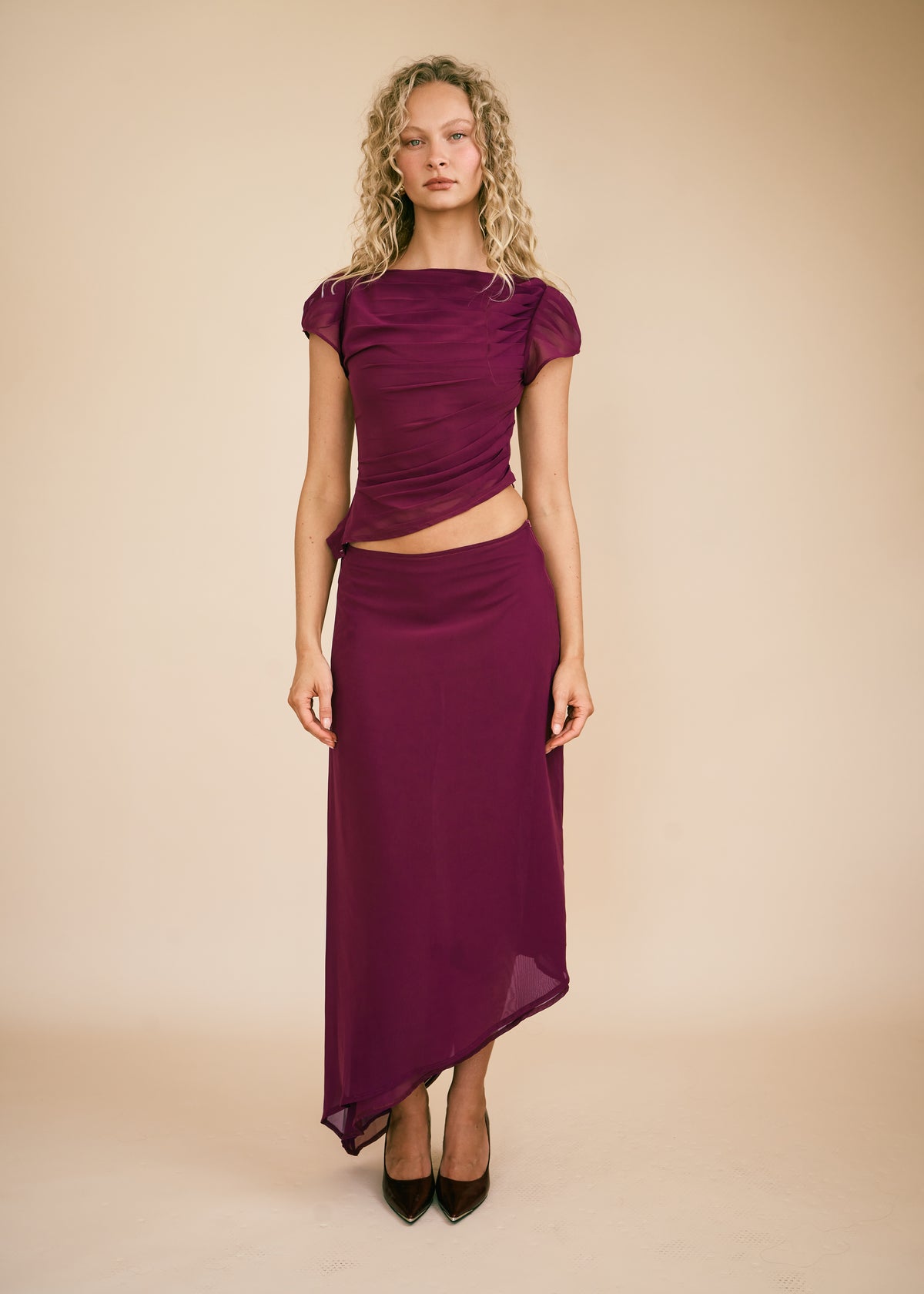 A woman modeling a deep plum purple two-piece set consisting of a short-sleeve top with a twisted waist detail and a matching high-waisted draped skirt, paired with black pointed-toe heels against a solid beige studio backdrop.