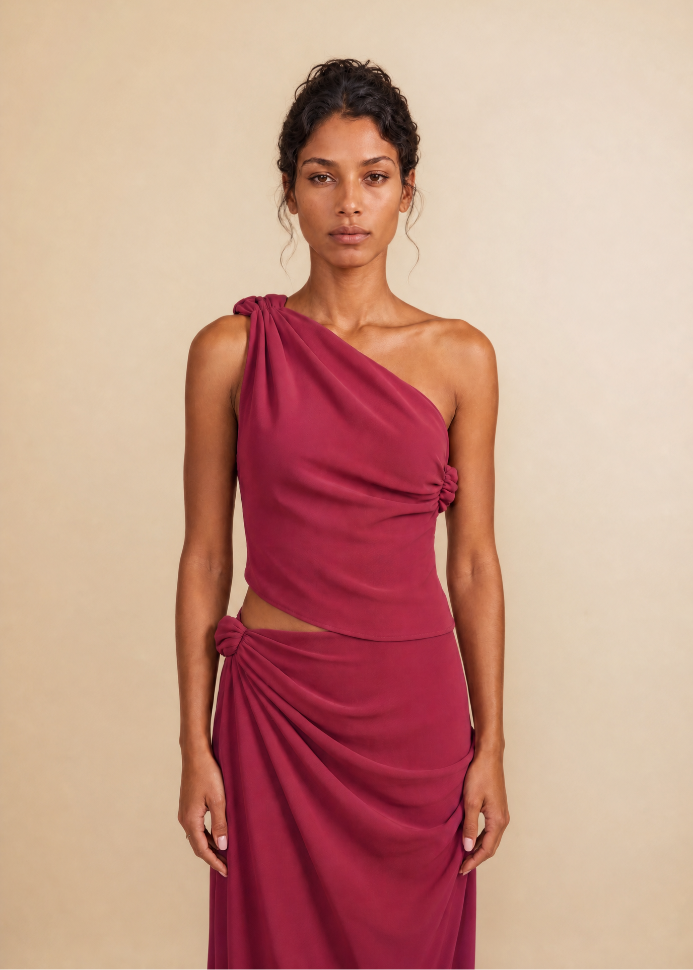 A woman modeling a one-shoulder draped dress in a deep magenta color, standing against a solid beige background.