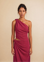 A woman modeling a one-shoulder draped dress in a deep magenta color, standing against a solid beige background.
