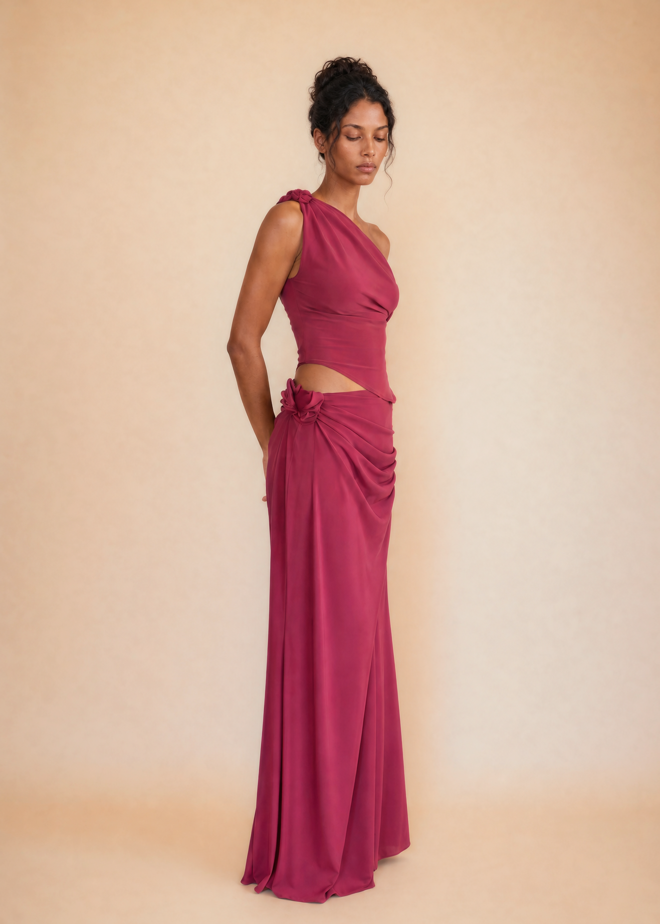 A woman modeling a floor-length magenta gown with a one-shoulder neckline, a midriff cutout, and gathered draping detail at the waist, photographed against a warm beige studio backdrop.