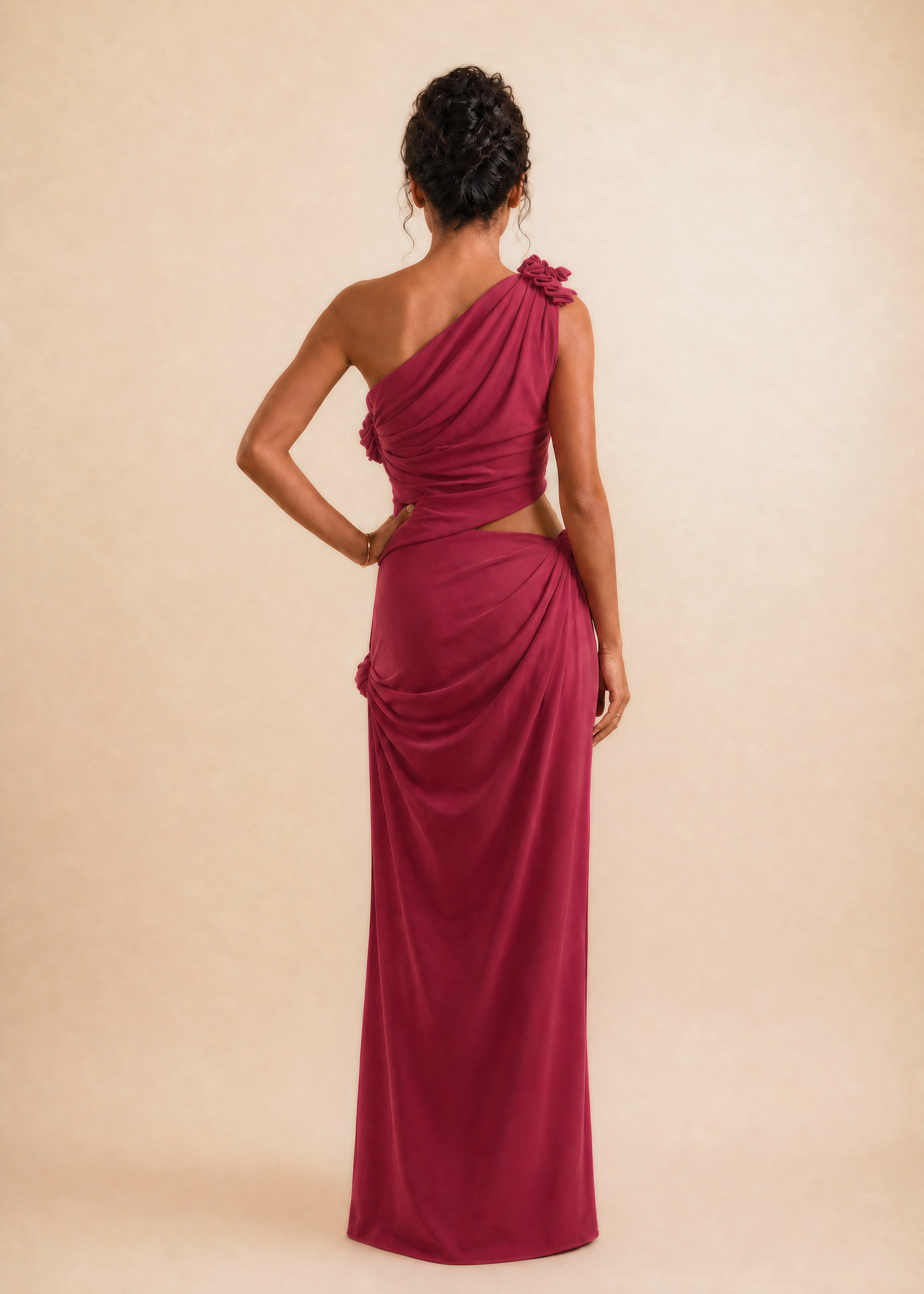 A woman modeling a floor-length, one-shoulder magenta dress from the back, standing against a solid beige studio backdrop.