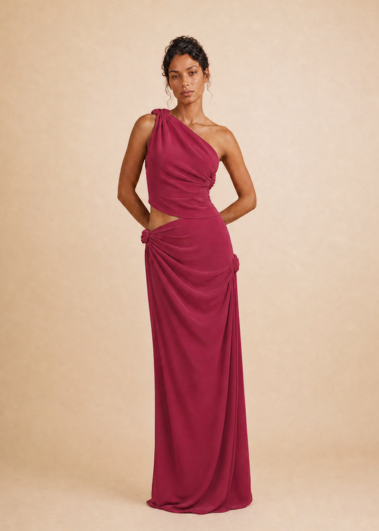 A woman modeling a floor-length, one-shoulder draped gown in a rich magenta-pink color, standing against a solid beige studio backdrop.