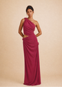 A woman modeling a floor-length, one-shoulder draped gown in a rich magenta-pink color, standing against a solid beige studio backdrop.