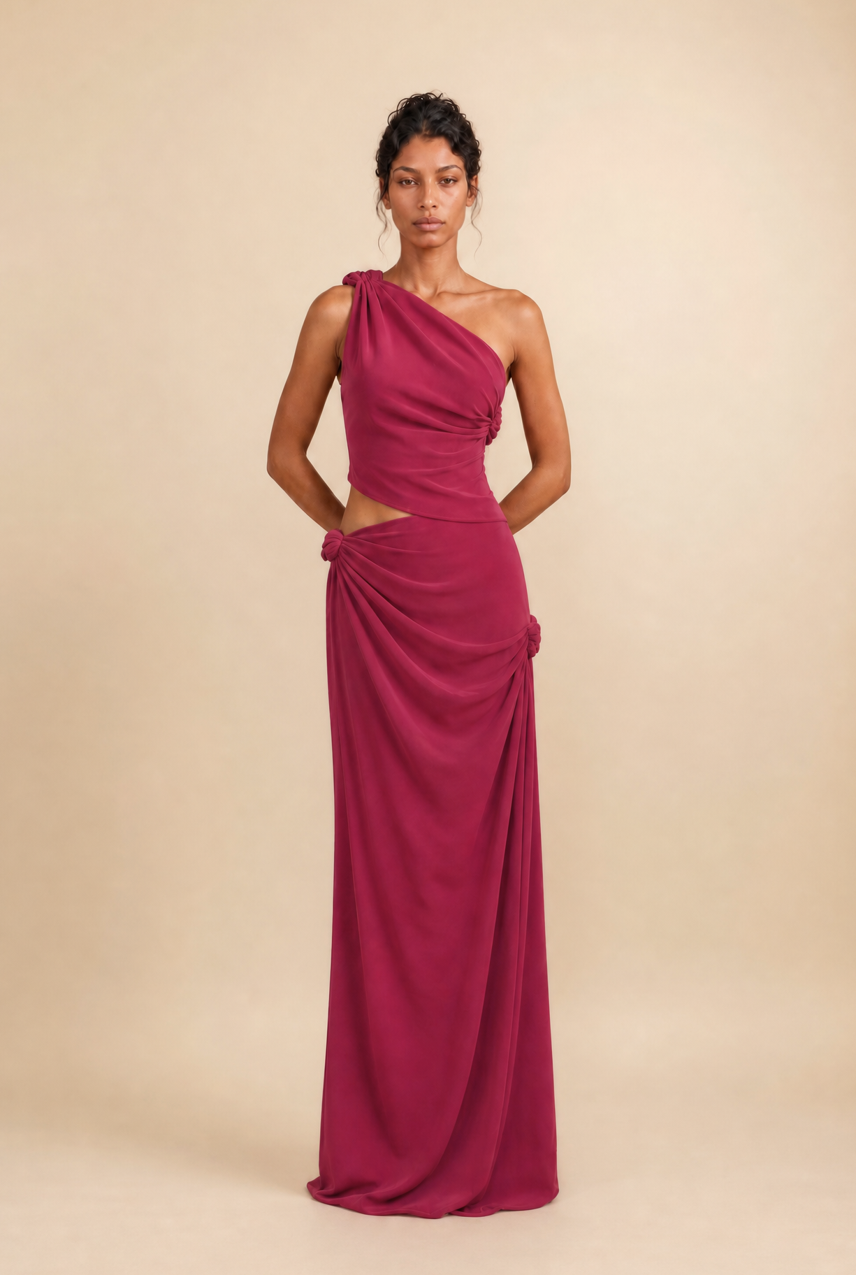 A woman modeling a floor-length magenta one-shoulder gown with a draped, gathered skirt against a plain beige studio background.