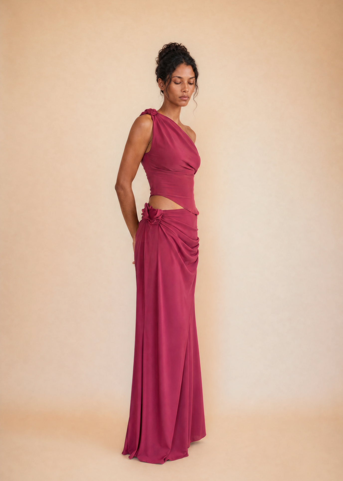 A woman modeling a floor-length magenta gown with a one-shoulder neckline, a midriff cutout, and gathered draping detail at the waist, photographed against a warm beige studio backdrop.